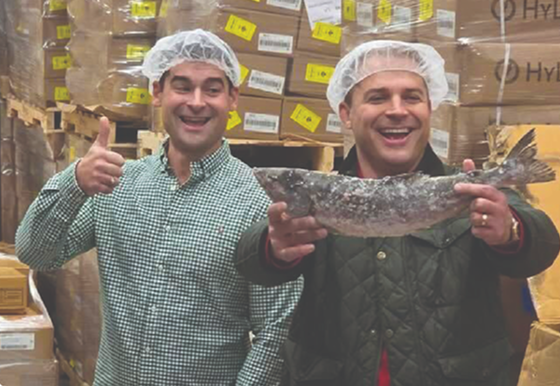 Two men wearing white hairnets stand in a warehouse holding a large whole salmon. One man on the left wears a green and white checkered shirt and gives a thumbs-up, while the man on the right wears a dark quilted jacket and holds the salmon horizontally.