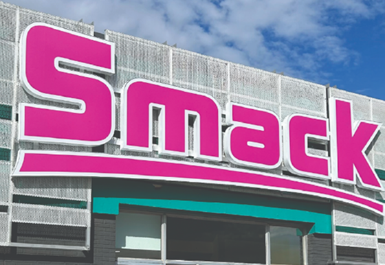 A large pink and white 'Smack' logo sign mounted on the exterior of a building with a gray textured facade and a teal awning above the entrance.