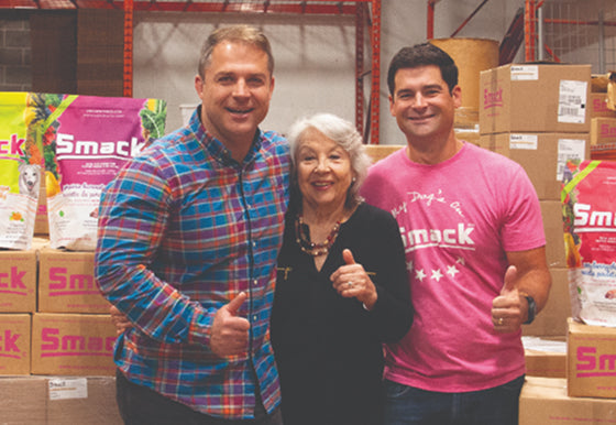 Three people standing together in a warehouse or storage area with boxes and Smack-branded bags visible in the background. The person on the left wears a blue and red plaid shirt, the center person wears black, and the person on the right wears a bright pink Smack T-shirt.